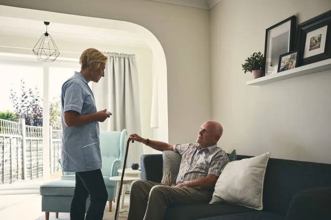 Ein älterer Patient mit Krückstock sitzt daheim auf der Couch. Vor ihm steht eine Pflegerin und spricht mit ihm. 